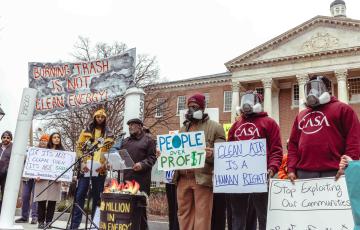 People rallying in front of the MD State House, some wearing gas masks, with signs like "Burning trash is not clean energy," "clean air is a human right," and "people over profit."