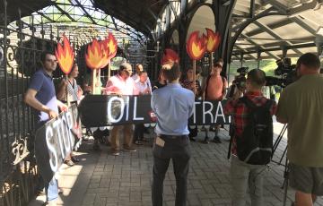 15 people hold signs saying "Stop Oil Trains" and showing train tank cars and flames as news media films