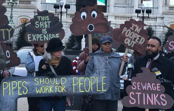 A crowd of people rallying outside of Baltimore City Hall, with signs like "Pipes should work for people" and "sewage stinks"